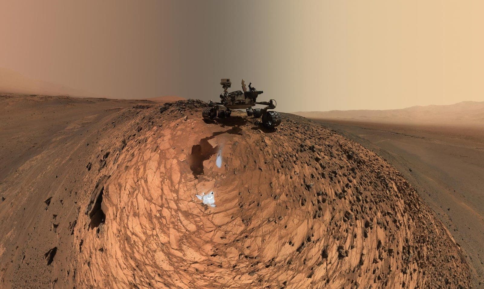 Curiosity seen from ground level on top of a rounded, rock-strewn rise, with two pale drill patches on the reddish stone below it.