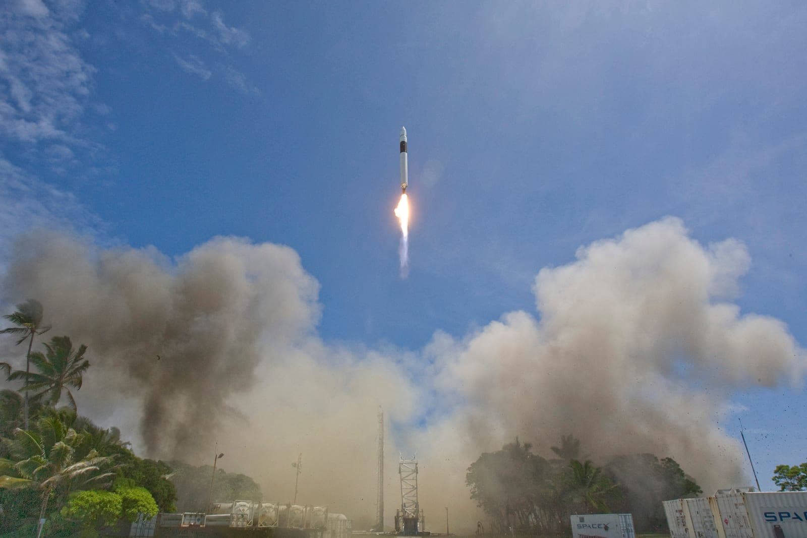 A slim white rocket with a black band climbs on a bright flame into a blue sky, above a small launch tower half hidden in billowing dust, with palm trees at the left.