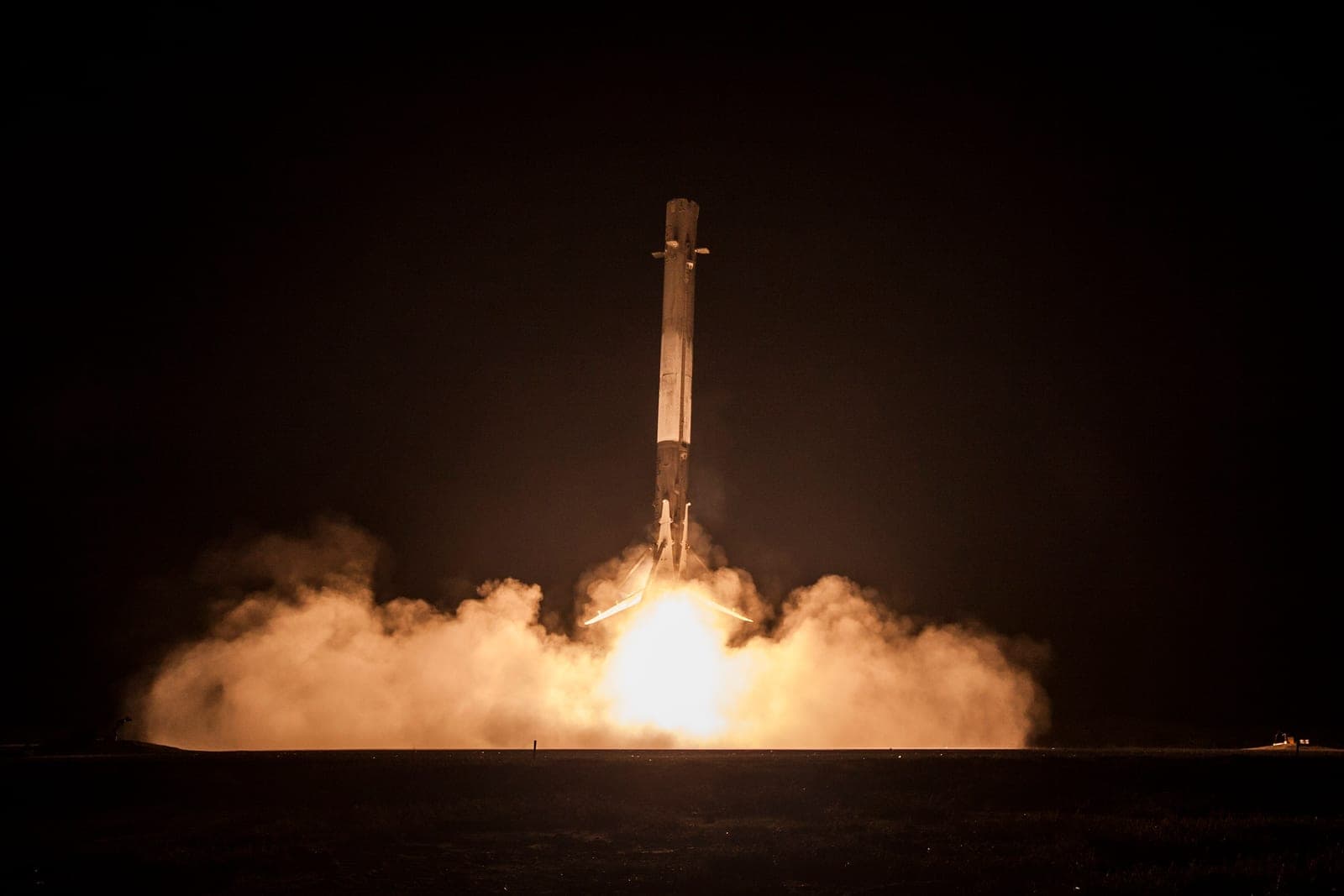 At night, a tall rocket stage descends tail-first onto flat ground on a bright engine flame, landing legs spread beneath it and a glowing cloud of exhaust spreading out on both sides.