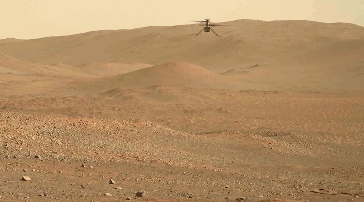 A wide Martian landscape of low dunes and scattered rock, with a small four-rotor helicopter airborne above the ridge line.