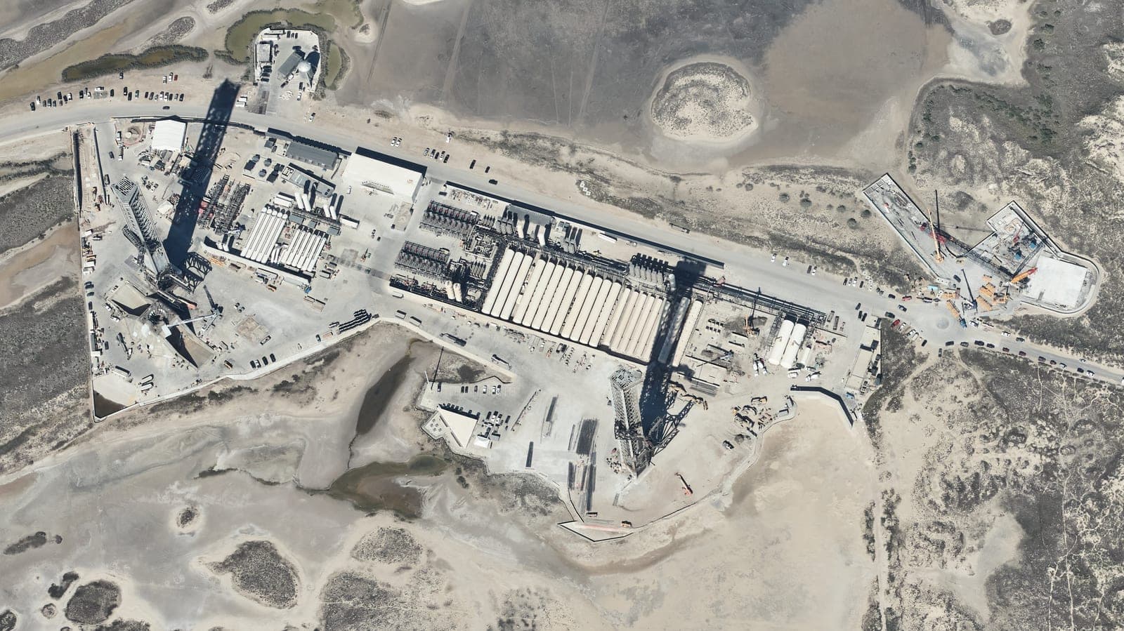 An aerial photograph of a coastal launch site on flat sand and scrub: two tall launch towers casting long shadows, rows of large white storage tanks, buildings and parked cars along a road.